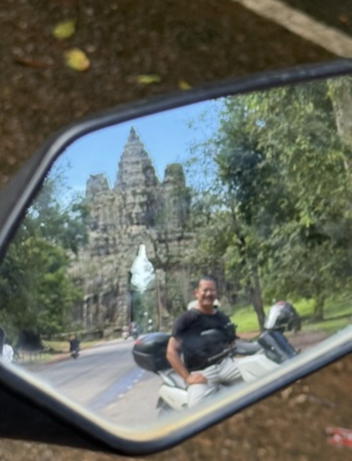 Boramy on a motorbike reflected in a side mirror with the south gate of Angkor Thom rising behind him, Cambodia