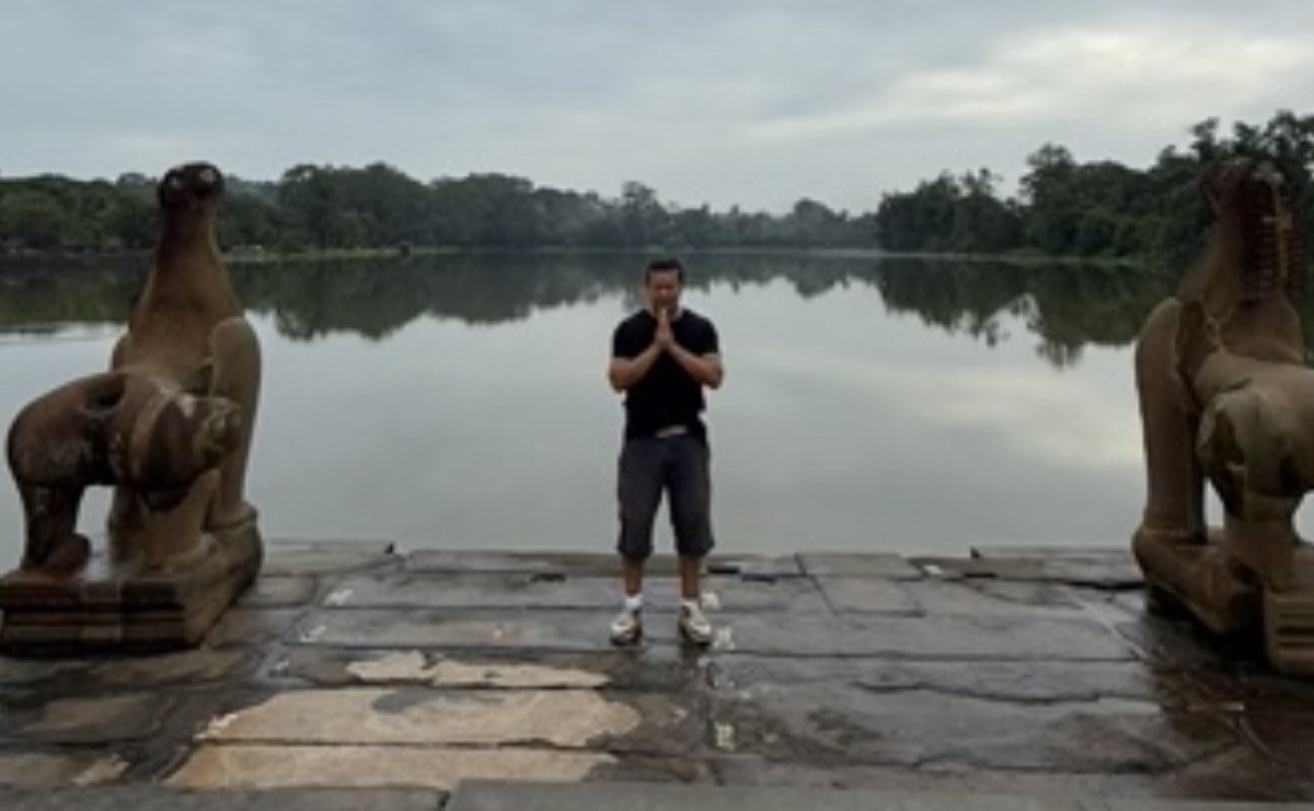 Boramy in prayer at dawn between ancient guardian statues at the water's edge, Angkor, Cambodia