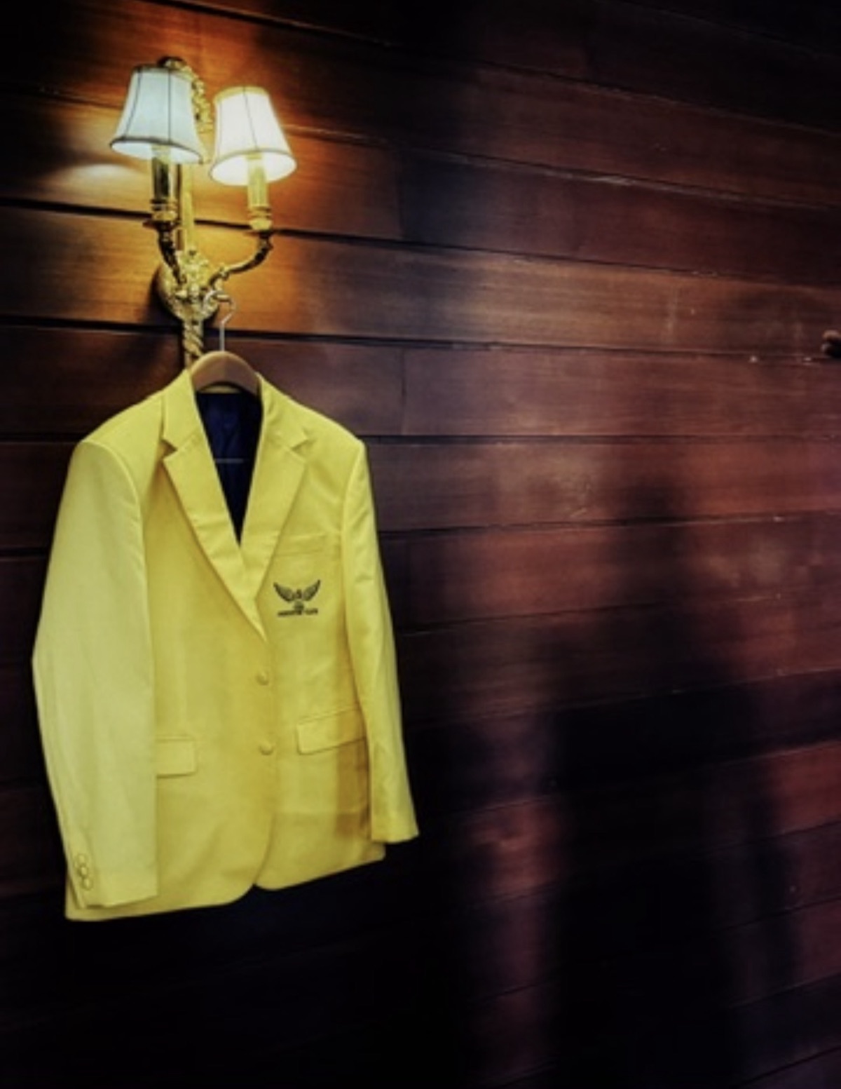 The first physical Country C1ub gold jacket — one of one, with the gold wing crest — hanging from a brass sconce against dark wood