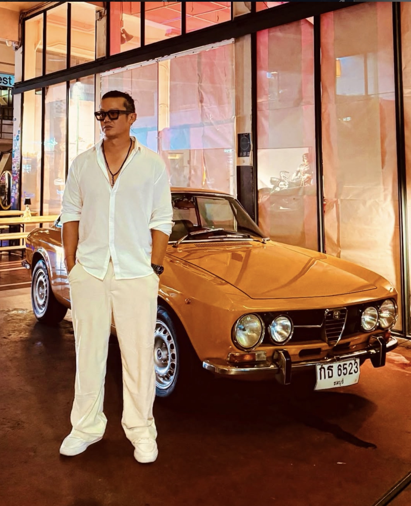 DJ Boramy in white linen beside a vintage Alfa Romeo on a neon-lit night in Southeast Asia